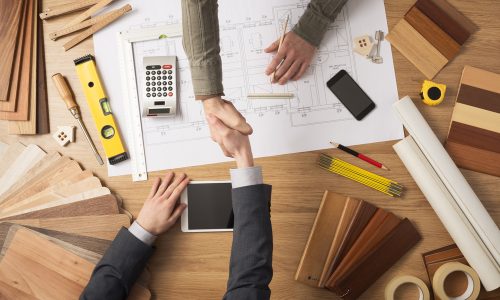 Architect and customer businessman shaking hands top view, desktop with building project, tools and wood swatches on background